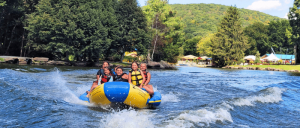 Four people enjoying a fun ride on a yellow and blue inflatable boat, splashing through a river surrounded by lush greenery.