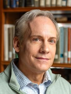 Close-up headshot of a middle-aged person with short gray hair and light-colored eyes, wearing a light green sweater over a checkered shirt, seated in front of bookshelves in a softly lit library setting