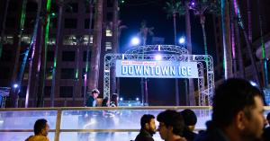 Image of the San Jose Downtown Ice Rink, a circular rink surrounded by palm trees, with warmly dressed ice skaters skating under a sign that reads "back by popular demand: downtown ice."