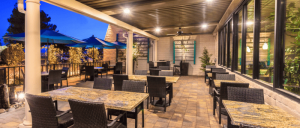 An outdoor dining area featuring dark wicker tables and chairs, with umbrellas, surrounded by greenery at dusk.