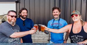 Exterior of Mare Island Brewing Co.’s Coal Shed Brewery with smiling guests raising pints of beer on the Mare Island Waterfront in Vallejo, CA.