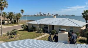 Rooftop view of a coastal suburban neighborhood with a metal-roofed house, palm trees, and distant city skyline across the bay under a clear blue sky.