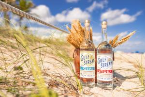 Gulf Stream Distillery vodka and gin bottles displayed on a Fort Lauderdale beach with sea oats and coastal rope, showcasing Florida-crafted, gluten-free spirits inspired by coastal living.