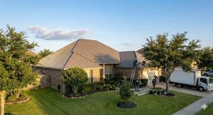 Aerial view of a suburban brick house with a metal roof. Workers are on ladders near the roof, with tools and a truck nearby. The sky is clear, creating a calm and industrious atmosphere.
