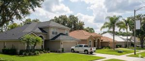 Suburban street with modern houses, lush green lawns, and a parked white truck. Palm trees line the sidewalk under a bright, partly cloudy sky. Peaceful scene.