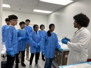 A group of students wearing blue lab coats, safety goggles, and gloves stand around a lab bench while an instructor in a white lab coat explains a laboratory procedure in a biomedical classroom setting.”