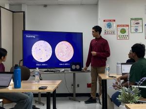 A student stands beside a large screen displaying microscope images of stained samples while presenting to classmates seated around lab tables in a science classroom.
