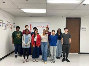 A group of students stand in two rows inside a classroom, smiling in front of an Alamo BioCenter banner that reads ‘Lab of Limitless Discoveries,’ with science posters visible on the walls.