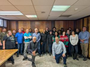 Group photo of Oakwood Veneer employees standing and kneeling together in an office showroom, with a wall display of various wood veneer panels behind them, celebrating the company’s 40 years in business.