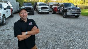 RJR Demolition & Junk founder Ryan Gibbons standing with arms crossed in front of a fleet of branded work trucks and dump trailers used for junk removal and demolition services.