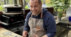 A smiling man wearing an apron gives a thumbs-up while standing behind a freshly baked pizza on a cooling rack at an outdoor cooking station.