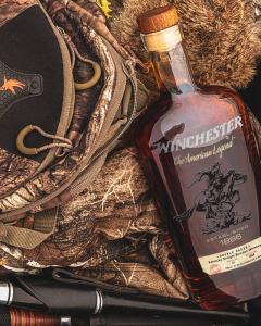 A bottle of Winchester Double Oaked Bourbon resting on camouflage hunting gear, emphasizing rugged outdoor heritage and American craftsmanship.