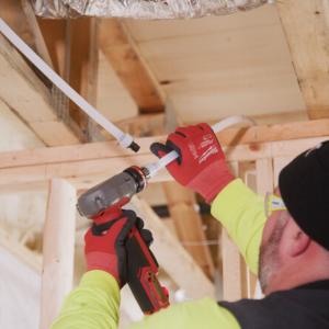 A close-up of a technician in red safety gloves using a power tool to install white PEX piping into a wooden ceiling frame.