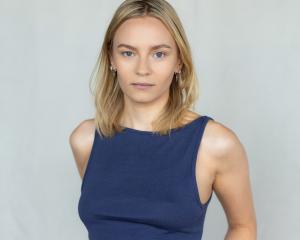 Professional studio headshot of a female actor with blonde hair and blue eyes, wearing a navy sleeveless top, photographed against a neutral light background.
