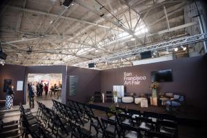 Wide interior view of the San Francisco Art Fair at Fort Mason Festival Pavilion showing the expansive exhibition layout, structured seating area, stage setup, overhead truss lighting, and overall spatial proportion of the fair.