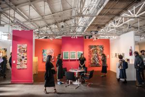 Color rich gallery booths, contemporary artworks on display, visible overhead truss structures, and a spacious, well defined exhibition layout.