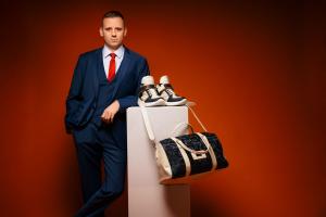 A portrait of Lian Ospelt, founder of Lian Original, wearing a tailored navy three-piece suit and red tie. He is leaning against a white pedestal that displays a pair of high-top sneakers and a camouflage-patterned travel bag, symbolizing the brand's fusi