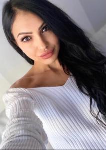 Portrait of Giulia Deamina wearing a white off-the-shoulder top, with long dark hair and a neutral background, photographed in a clean, professional style.