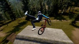 Third-person view of a mountain biker performing a trick above a wooden ramp in a forested environment. Motion blur emphasizes the rider's speed, with green hills and trees in the background