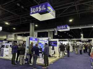 Image set in the indoor area of a large international trade fair, featuring wide aisles covered with blue carpet. At the center of the scene, the Brazil Pavilion stands out, identified by suspended ceiling cubes displaying the word “Brasil” in white lette