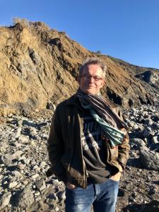 Author Alan Collenette stands on a rocky coastal beach beneath a sunlit cliff, wearing glasses, a jacket, jeans, and a scarf.