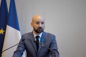 Official photograph of a senior economic and financial expert delivering a formal address at a lectern, wearing the insignia of Knight of the French National Order of Merit. Set against a neutral institutional backdrop with French and European flags, the