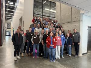 A group of approximately 40 Sleep in Heavenly Peace volunteers posed together on and around a cement staircase in a bright technical learning center with floor-to-ceiling windows. The group includes men and women of various ages wearing casual clothing an