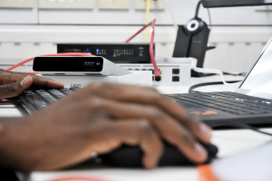 Close-up of a technician's hands typing on a keyboard, with a Ubiquiti Cloud Key and SonicWall firewall visible in the background, representing IT infrastructure management.