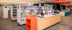 Modern library interior featuring bookshelves, a checkout counter, and an information desk, with bright lighting and an orange cart.