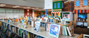 A bright library interior featuring shelves of colorful books and a playful mural of a town in the background.