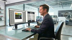 Car dealership employee seated at a desk editing digital signage content on dual computer monitors.