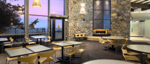 Modern dining area with stone walls, tables, and chairs, illuminated by pendant lights, overlooking a scenic sunset.
