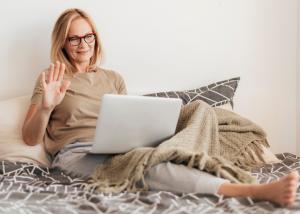 Woman sitting comfortably on couch with laptop waving during virtual therapy session, representing accessible telehealth mental health services