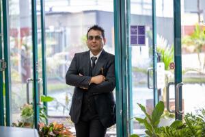 A professional portrait of Maddy Gupta, founder and CEO of Manhari Recycling, wearing a dark suit and standing with arms crossed in a bright, modern office doorway with greenery in the background.