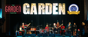 A live band performs on stage at the Garden Theater, with festive decorations and a "Certified Autism Center" badge displayed.