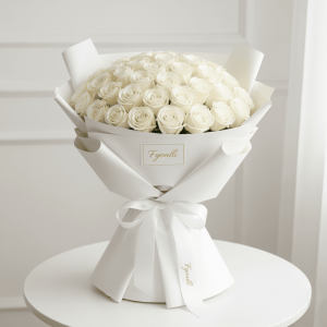 A bouquet of elegant white roses wrapped in smooth white paper, tied with a ribbon, sits on a round table against a soft white background, exuding elegance.