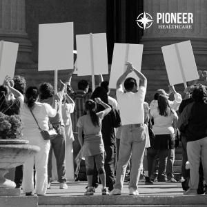 A powerful black and white photograph captures healthcare workers from behind as they stand on outdoor steps during a protest or demonstration. Multiple participants hold white signs raised above their heads, creating a striking visual representation of c
