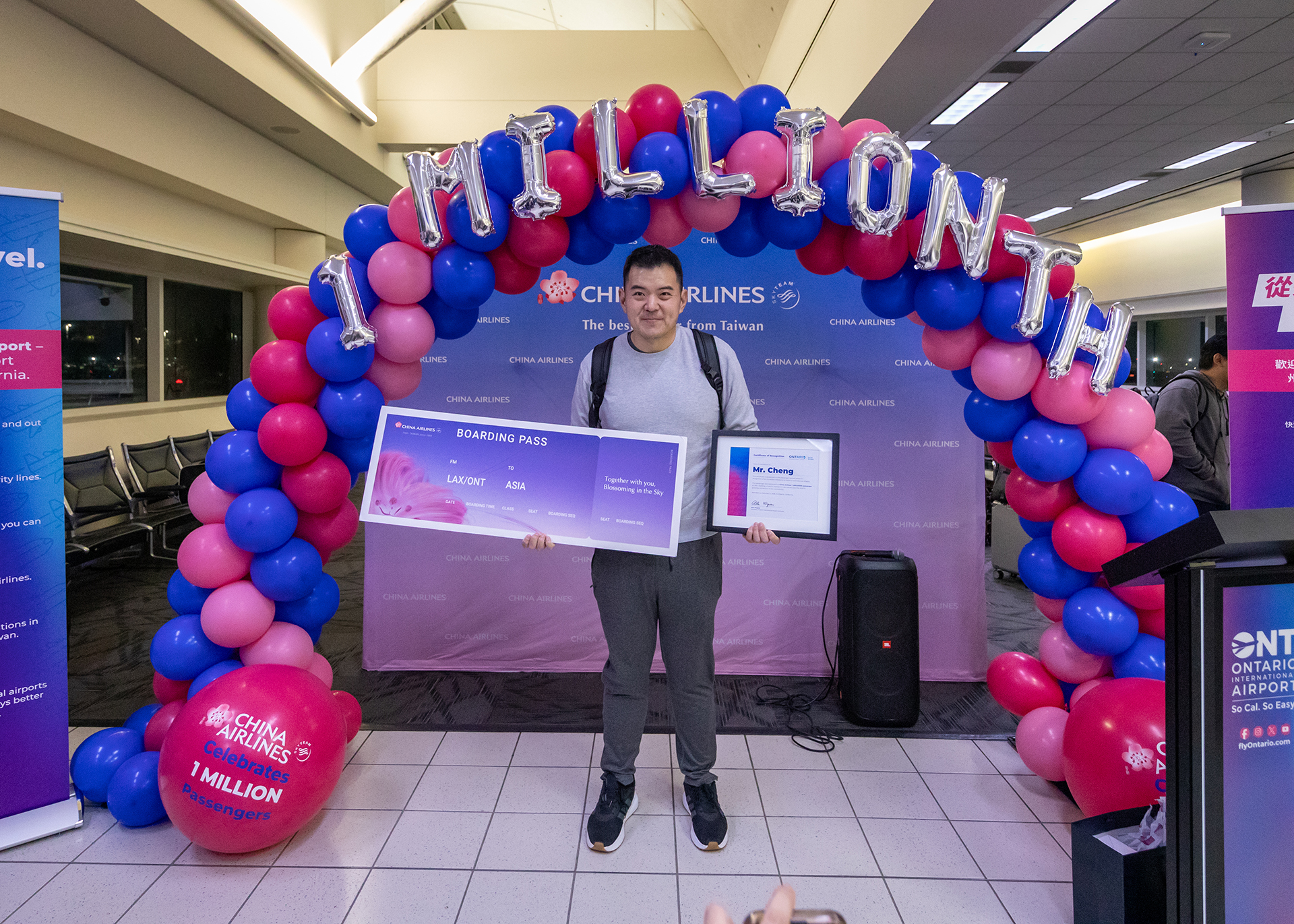 China Airlines’ 1 millionth passenger, Mr. Cheng, stands beneath a balloon arch at Ontario International Airport holding a commemorative boarding pass and certificate after winning a roundtrip ticket to Asia.