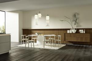 A spacious, modern dining room with a white circular table, light wood chairs, and a long walnut sideboard against a matching wood-slat wall. Three sculptural white pendant lights hang above.