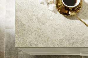 A high-angle detail shot of a light-colored, marbled countertop. A gold saucer with a cup of black coffee and a gold spoon are visible in the top right corner.
