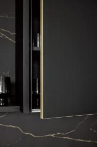 A close-up of a black cabinet door slightly open, highlighting the thin, brushed brass-colored finish on the vertical edge of the door.