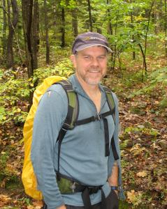 Peter Palmiotto smiling on a wooded trail, wearing a cap, long-sleeve shirt, and backpack.