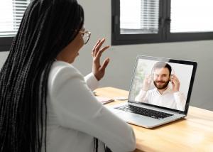 A woman seated at a desk waves during a video call with a healthcare provider on a laptop, representing a virtual mental health or telehealth appointment.