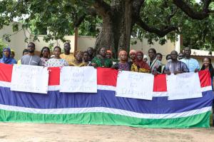 A group of 23 people in The Gambia stand roughly in line under a large tree, with four hand written signs saying "Promoting FGM is Illegal", "FGM is a human rights violation", "FGM is Illegal",  "FGM is not a religious obligation"