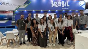 La imagen muestra un grupo de personas sonriendo y posando para una foto frente al stand de Brasil en Colombiatex 2026, un evento internacional. El grupo está compuesto por hombres y mujeres de diferentes edades y etnias, todos vistiendo camisetas blancas