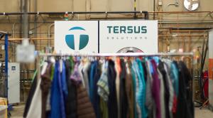 Image of a Tersus CO2 cleaning machine (10'x10' white cube with a vault-like door on the front)with a rack of resale clothing in front of it.
