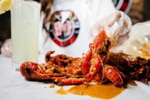 Close-up of a gloved hand holding a seasoned crawfish over a table covered in saucy crawfish shells, with a cup of lemonade on the side.