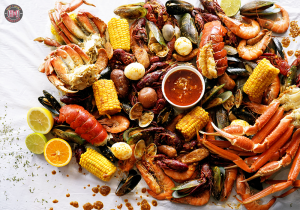 Overhead view of a large seafood boil spread across a table, featuring crab legs, crawfish, shrimp, lobster tails, mussels, clams, corn on the cob, potatoes, sausage, boiled eggs, lemon slices, and a cup of dipping sauce in the center.