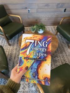 Hand holding 7 ING energy Types book by Stephanie Mason in cozy setting with green chairs and wood paneling