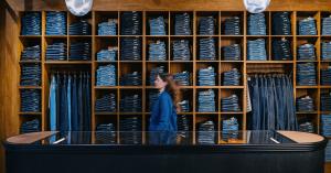 Intérieur de la boutique Nudie Jeans à Bristol, avec un mur d’étagères en bois rempli de jeans pliés et suspendus, et une personne passant devant le comptoir.